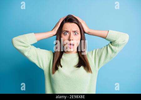 Foto von schockiert Angst niedlichen Mädchen öffnen Mund Hände Kopf Tragen Sie grünen Pullover isoliert blaue Farbe Hintergrund Stockfoto