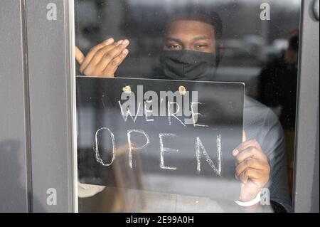 Ein junger afrikanischer Mann ist Café-Manager, Besitzer in schützende medizinische Maske hält offen Schild breit, Blick durch Glastür. Wiedereröffnung von Kleinunternehmen Stockfoto