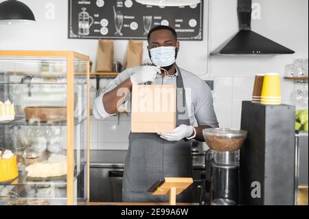Ein afroamerikanischer Kellner trägt Gesichtsmaske und Handschuhe Bestellung zum Mitnehmen im Öko-Paket in den Händen, die am stehen Bar-Theke in gemütlichen Kaffee Stockfoto