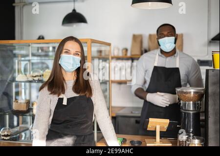 Vielfältiges Team im Cafe. Multirassische Kellner und Kellnerin in Schürzen, tragen medizinische Masken und Handschuhe stehen in einem gemütlichen Café, Sicherheit während Pandemie-Periode Stockfoto