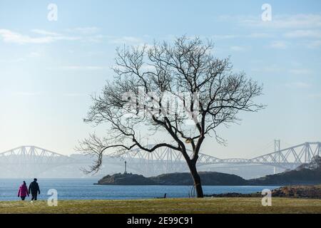 Ein Paar, das den Fife Coastal Path entlang in Dalgety Bay, Fife, mit der Forth Bridge in der Ferne geht. Stockfoto