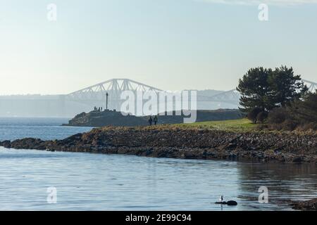 Ein Paar, das den Fife Coastal Path entlang in Dalgety Bay, Fife, mit der Forth Bridge in der Ferne geht. Stockfoto