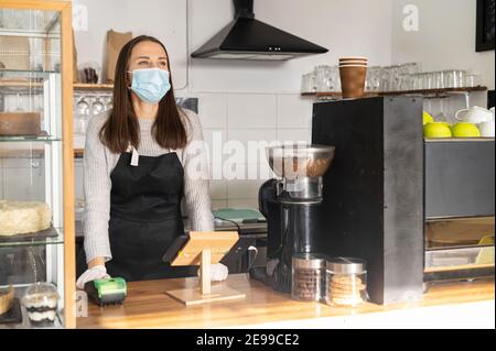 Hinter der Arbeitsplatte steht eine maskentragende Cafébesitzerin und freut sich auf die Kunden. Eine Kellnerin im Vorfeld in der Bäckerei während Pandemie und Stockfoto