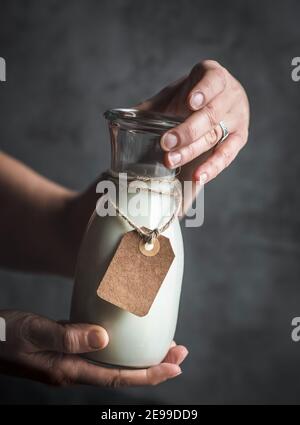 Weibliche Hände halten Flasche Mandelmilch mit leerem Papieretikett. Dunkel und launisch. Speicherplatz kopieren. Stockfoto