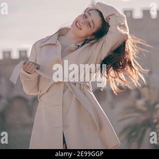 Oortrait einer schönen jungen Frau auf den Straßen von Valencia in einer braunen Gabardine an einem sonnigen Tag bei Sonnenuntergang posiert und lächelt. Mode Saisonal 2021. Stockfoto