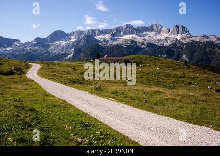 Leere Schotterstraße in den Alpen mit Blick auf die Berge von Canin in der Ferne und grüne Weiden mit Scheune in der Mitte. Stockfoto