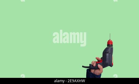 Elektrische Bohrmaschine in der Hand auf grünem Hintergrund, Bauwerkzeug. Ein Mann im Bauüberbau hält einen Bohrer in der Hand. Stockfoto