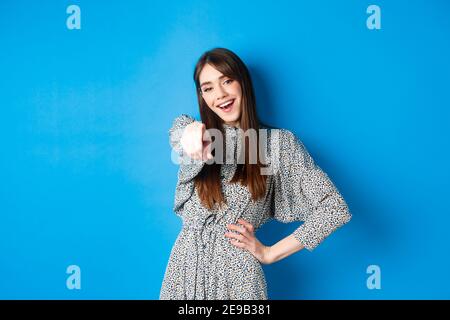 Motivierte und aufgeregte junge Frau, die mit dem Finger auf die Kamera zeigt und lächelt, Sie gratuliert, lobt oder lädt, glücklich auf blauem Hintergrund steht Stockfoto