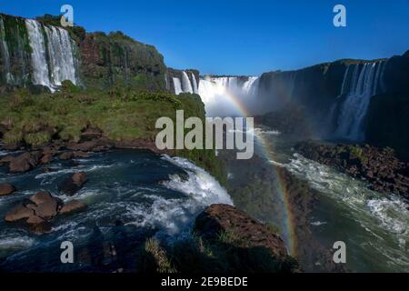 Ein Blick auf die Iguazu-Wasserfälle in Brasilien, die in weiter Ferne auf den Hals des Teufels schauen. Im Vordergrund hat sich ein Regenbogen gebildet. Stockfoto