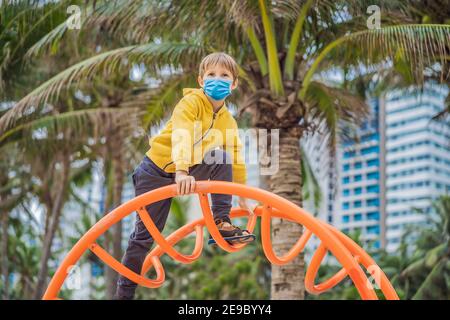 Der Junge trägt eine medizinische Maske während des COVID-19 Coronavirus klettert Auf dem Spielplatz vor dem Hintergrund von Palmen Stockfoto