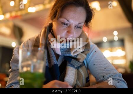 Junge Frau trägt Schal mit Getränk in hellen bunten Cocktail Bar mit Handy Blick auf den Bildschirm Beleuchtung ihr Gesicht vom Telefon Stockfoto