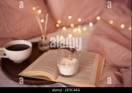 Brennende Kerze bleibt auf offenem Papierbuch und Tasse Tee mit Duftstäbchen im Hintergrund über glühenden Lichtern. Gemütliches Atmosphärenkonzept. Guten Morgen. H Stockfoto