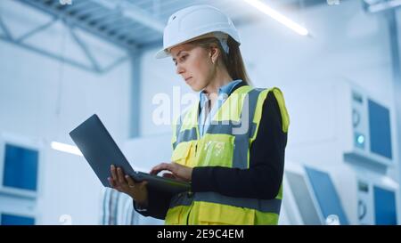 High-Tech-Fabrik: Selbstbewusste und professionelle weibliche Ingenieurin trägt Sicherheitsjacke und Harthut Halten und Arbeiten auf Laptop-Computer. Modern Hell Stockfoto