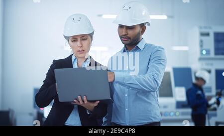 Weibliche leitende Ingenieurin und männliche Projektmanager stehen in der Mitte der modernen Fabrik, Laptop-Computer verwenden, Produktionsoptimierung planen Stockfoto