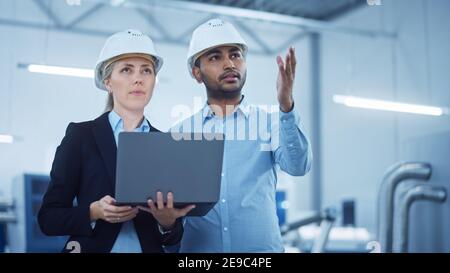 Weibliche leitende Ingenieurin und männliche Projektmanager stehen in der Mitte der modernen Fabrik, Laptop-Computer verwenden, Produktionsoptimierung planen Stockfoto