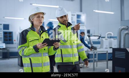 Chefingenieur und Projektleiter in Sicherheitswesten und Hartmücken gehen durch die moderne Fabrik, sprechen, Produktion optimieren. Industrieanlage Stockfoto