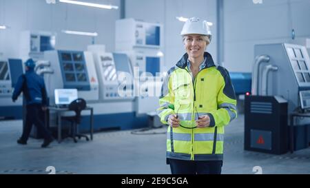 Schöne lächelnde weibliche Ingenieur trägt Sicherheitsweste und HardHat hält Sicherheitsbrillen. Professionelle Frau, die in der modernen Fertigungsfabrik arbeitet Stockfoto