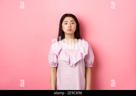 Traurig jammern asiatische Mädchen pouting und Stirnrunzeln, Blick aufgeregt über etwas unfair, beschweren, stehen in Kleid vor rosa Hintergrund Stockfoto