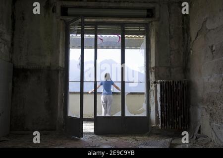 Frau, die auf einem Balkon in einem verlassenen Gebäude durch ein kaputtes Fenster gesehen steht. Stockfoto