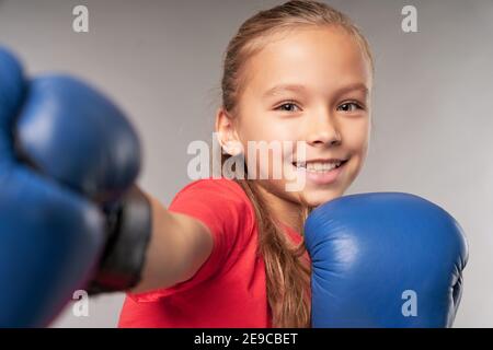 Liebenswert weibliches Kind in Boxhandschuhen Punching Luft und lächeln Beim Stehen vor grauem Hintergrund Stockfoto