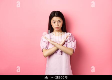 Ernsthafte chinesische Mädchen zeigen Kreuz Zeichen, machen Stop-Geste und sehen zuversichtlich, Block Person, sagen nein und lehnen Angebot, auf rosa Hintergrund stehen Stockfoto