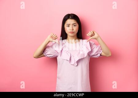 Skeptisch und enttäuscht asiatische Frau zeigt Daumen nach unten mit widerstrebenden Grinsen, nicht wie Produkt, zeigen negative Emotion, stehen auf rosa Hintergrund Stockfoto