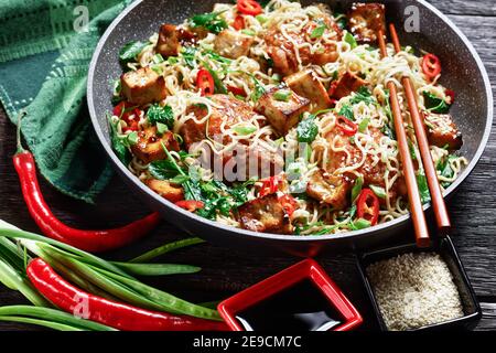 Süßer und saurer Tofu, Hühnerschenkel, chinesische Wok-Nudeln, wilder Grünkohl, Sesamsamen, rote Chili, grüne Zwiebel, serviert auf einer Pfanne mit Essstäbchen auf Stockfoto