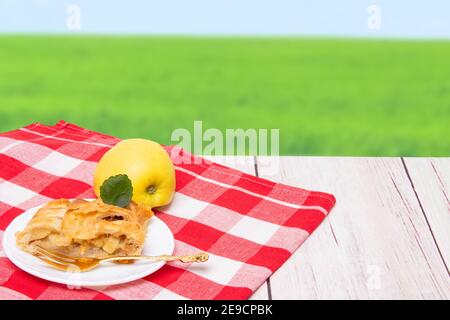 Apfelrolle serviert. Ein Stück hausgemachter frisch gebackener Apfelstrudel und ein frischer Apfel auf rotem Picknicktuch über unscharfem natürlichen Hintergrund. Osterfrühstückt Stockfoto