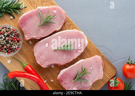 Rohes Schweinesteak mit Gewürzen, Rosmarin und Gemüse auf einem Holzbrett, zubereitete Fleischstücke auf grauem Betongrund Stockfoto