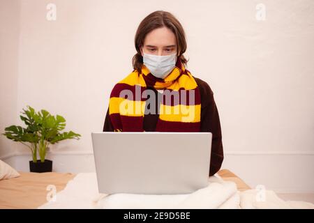 Ein kranker Mann sitzt mit Maske und Schal an einem Laptop. Coronavirus, covid, Home Quarantäne. Stockfoto