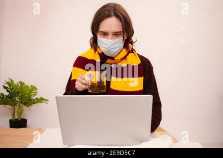 Ein kranker Mann sitzt mit Maske und Schal an einem Laptop. Coronavirus, covid, Home Quarantäne. Ein Mann trinkt Tee aus einer Erkältung. Stockfoto