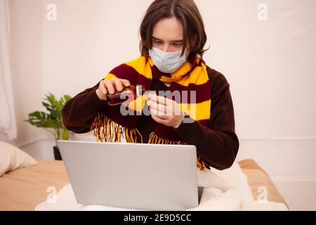 Ein kranker Mann sitzt mit Maske und Schal an einem Laptop. Coronavirus, covid, Home Quarantäne. Der Mann gießt sich Medikamente, behandelt Grippe, Erkältungen Stockfoto