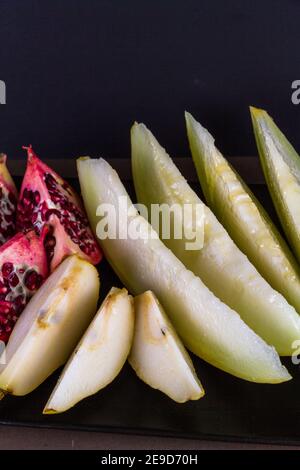 Granatapfel Melone und Birne in Scheiben geschnitten und auf Teller angeordnet. Dunkler Hintergrund, Porträt, Nahaufnahme, Makro Stockfoto