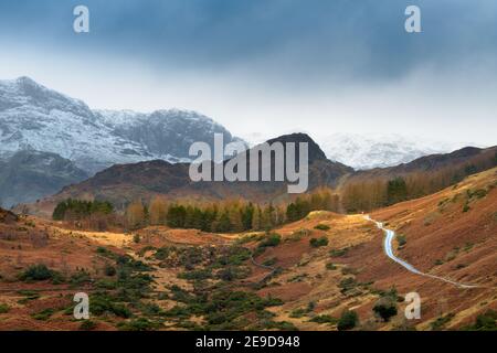 Einspurige Straße führt durch ländliche Landschaft mit schneebedeckten Bergen und dunklen Winterwolken. Langdale, Lake District, Großbritannien. Stockfoto