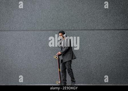 Moderner urbaner junger Dreadlocks Hipster Mann hält seinen Skate auf der Straße und hört Musik. Mit Kopfhörern auf dem Kopf gegen die schwarze Wand stehend. Stockfoto