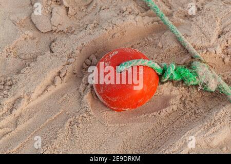 Grünes Fischernetz Seil mit rotem Schwimmer lag auf Küstensand. Nahaufnahme mit selektivem Fokus Stockfoto