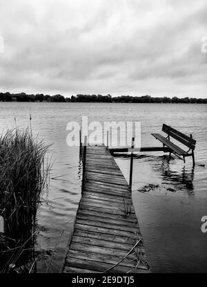 Vertikale Graustufen Aufnahme eines hölzernen Dock und Bank in der Nähe Ein See Stockfoto