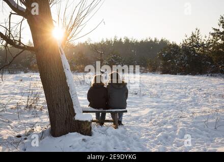 Junge Frau und ein Mann sitzen auf einer Bank mit dem Rücken im Rahmen in einem abgelegenen Ort, genießen einander und Winter Sonnenuntergang. Gute Beziehungen Stockfoto