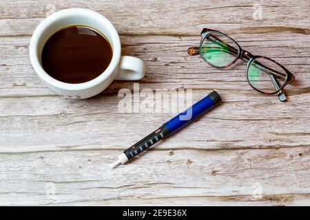 Ein Insulin-Stift für Diabetiker auf einem Holztisch oder Schreibtisch Neben einer Tasse Kaffee, um den Morgen darzustellen Injektion von Insulin Stockfoto
