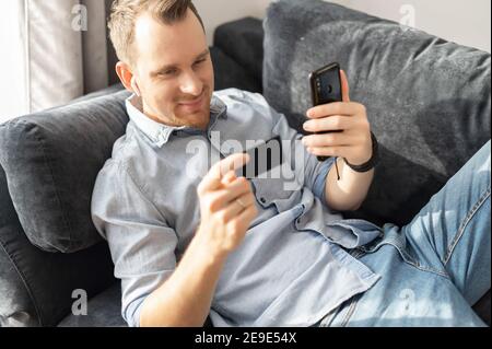 Ein junger Mann gibt Kreditkartendaten in das Smartphone ein, bezahlt und kauft online, während er zu Hause auf der bequemen Couch liegt Stockfoto