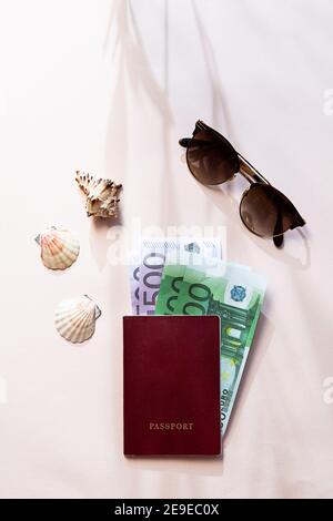 Reise Stillleben Szene mit Olivenbaum Ast Schatten im Sonnenlicht. Ein Pass mit Geld, Sonnenbrille und Muscheln, flach auf dem hellrosa Tisch Stockfoto