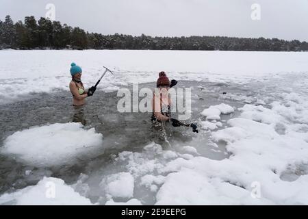 Aviemore, Schottland, Großbritannien. 4. Februar 2021. Loch Vaa ist völlig gefroren und bietet die Möglichkeit, über ihn zu laufen. Ein kleines Bootshaus ist nun eisgebunden und drei Damen aus der InVaa Dookers Wildschwimmgruppe nutzen Äxte, um das Eis zu brechen, damit sie für ein paar Minuten im eisigen Wasser einweichen können. PIC; Carolyn Stead und Wendy Cathcart brechen das Eis in Loch Vaa. Iain Masterton/Alamy Live News Stockfoto