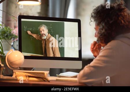 Rückansicht des jungen Mannes, der Computermonitor und ansieht Online lernen, während der Lehrer etwas sagt und auf die Tafel zeigt Stockfoto