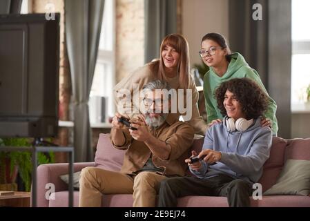 Vater und Sohn spielen Videospiel auf dem Sofa mit Tochter Und Mutter unterstützt sie im Hintergrund Stockfoto