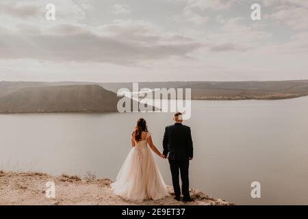 Liebevolles Paar Hochzeit Brautpaar in weißem Kleid und Anzug Spaziergang im Sommer auf dem Berg über dem Fluss. Sonnenuntergang und Sonnenaufgang. Mann und Frau auf Felsen über der Klippe Stockfoto