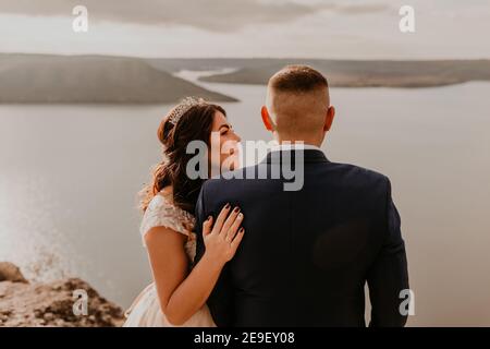 Liebevolles Paar Hochzeit Brautpaar in weißem Kleid und Anzug Spaziergang im Sommer auf dem Berg über dem Fluss. Sonnenuntergang und Sonnenaufgang. Mann und Frau auf Felsen über der Klippe Stockfoto