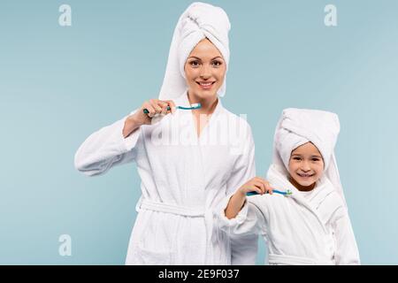 Lächelnde Mutter und Kind in Bademänteln mit Zahnbürsten und Zahnpasta Isoliert auf blau Stockfoto