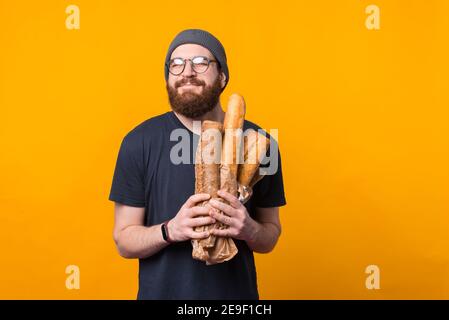 Foto eines bärtigen Hipster-Mannes, der frisch gebackene Brote auf gelbem Hintergrund hält. Stockfoto