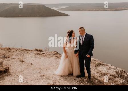 Liebevolles Paar Hochzeit Brautpaar in weißem Kleid und Anzug Spaziergang im Sommer auf dem Berg über dem Fluss. Sonnenuntergang und Sonnenaufgang. Mann und Frau auf Felsen über der Klippe Stockfoto