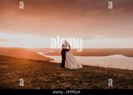 Ein liebevolles Paar Hochzeit Brautpaar Spaziergang im Sommer im Herbst auf Berg auf Klippe über dem Fluss. Sonnenuntergang. Braut in einem weißen Kleid mit Krone auf ihr Stockfoto